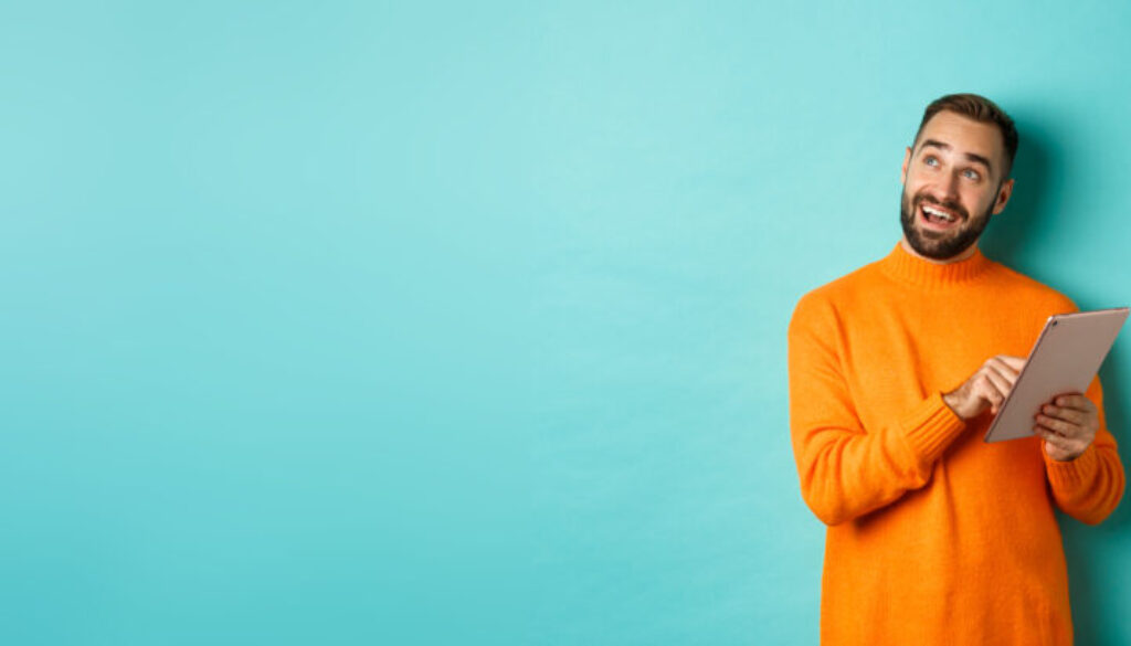 Handsome man thinking and using digital tablet, shopping online and pondering ideas, looking at upper left corner, standing over light blue background